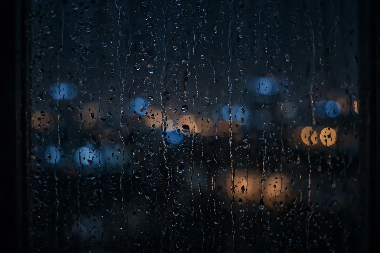 Rain on a dark window at night, city lights blurred beyond the glass